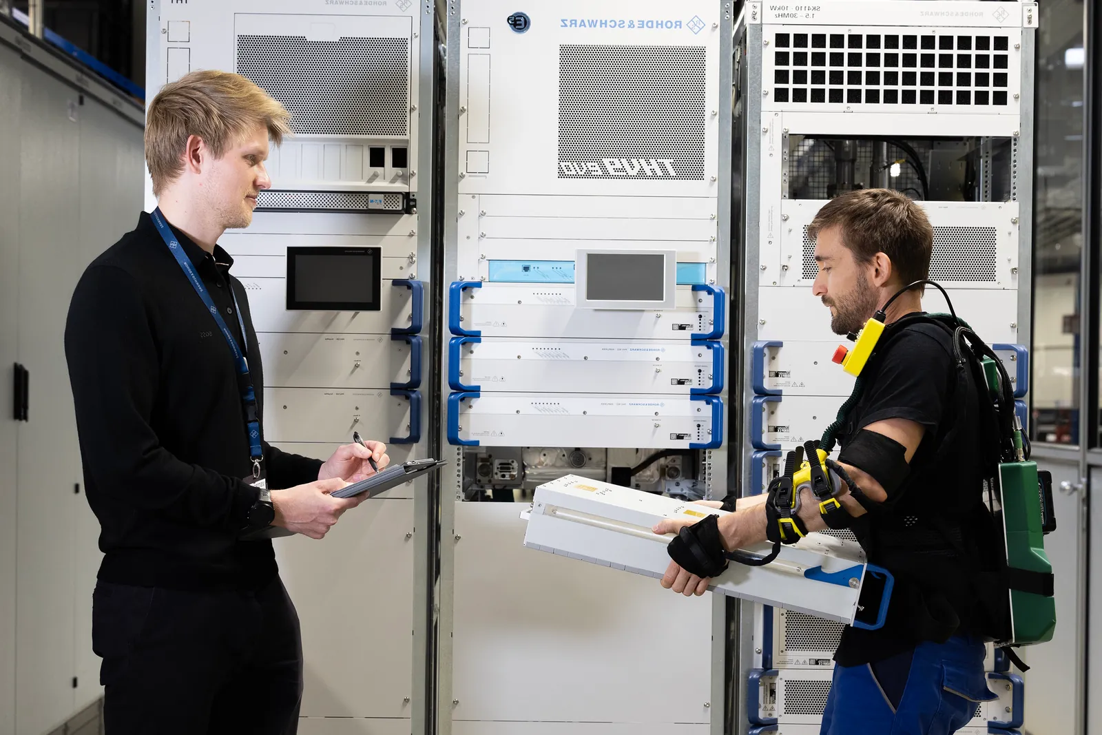 Ein Mitarbeiter testet das EASE Exoskelett im Werk von Rohde & Schwarz, begleitet von einem Beobachter mit Klemmbrett.